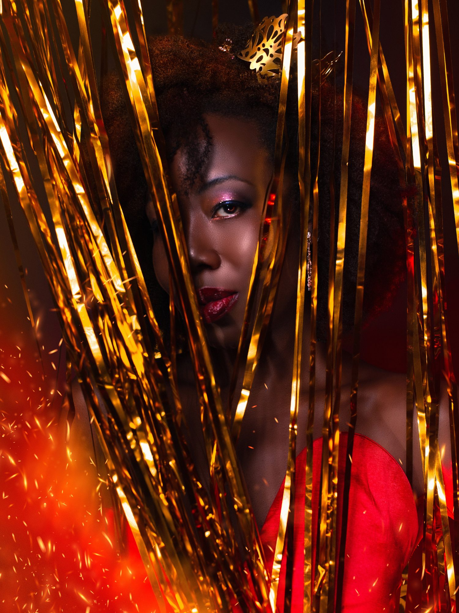 Radikal Queen in a Red Dress, Head and Shoulders, One Eye Peering Provocatively Through Gold Fringe Curtain.