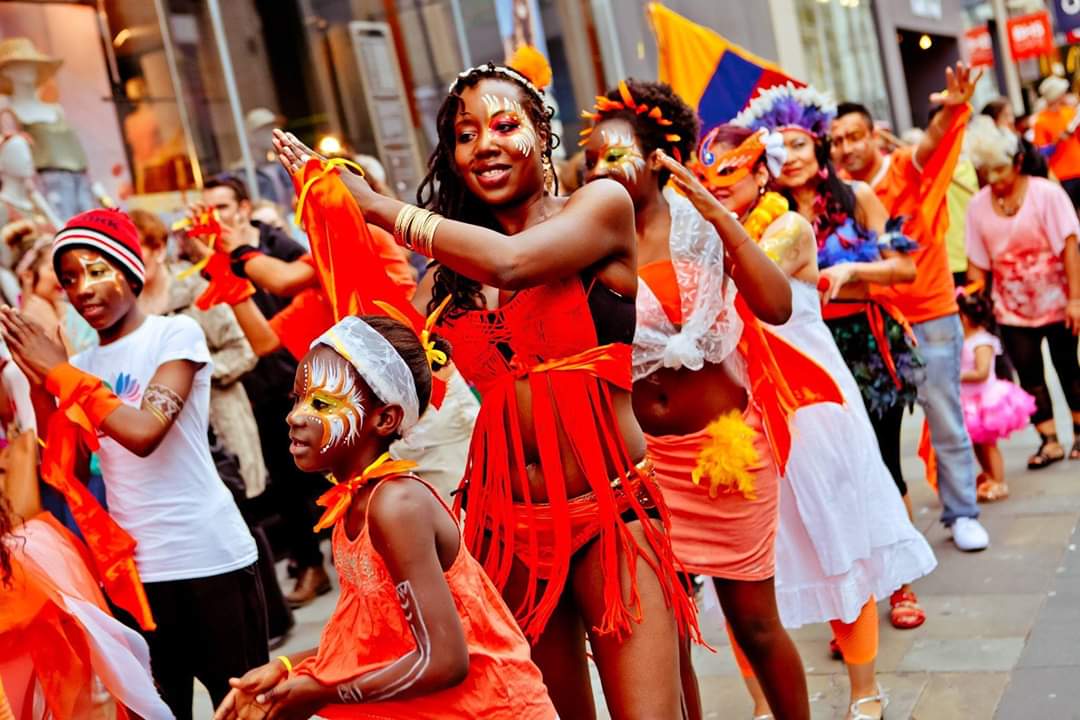 Radikal Queen in a Street Carnival with Several Black Children. All are Smiling, Dancing, Wearing Orange Costumes and Facepaint
